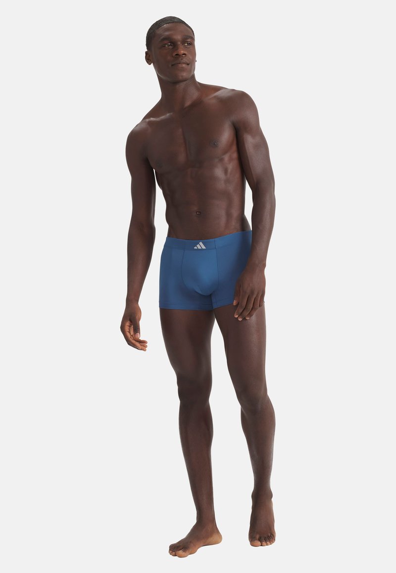Modèle masculin athlétique debout pieds nus, portant un boxer ajusté bleu avec un logo blanc sur la ceinture, sur un fond uni.