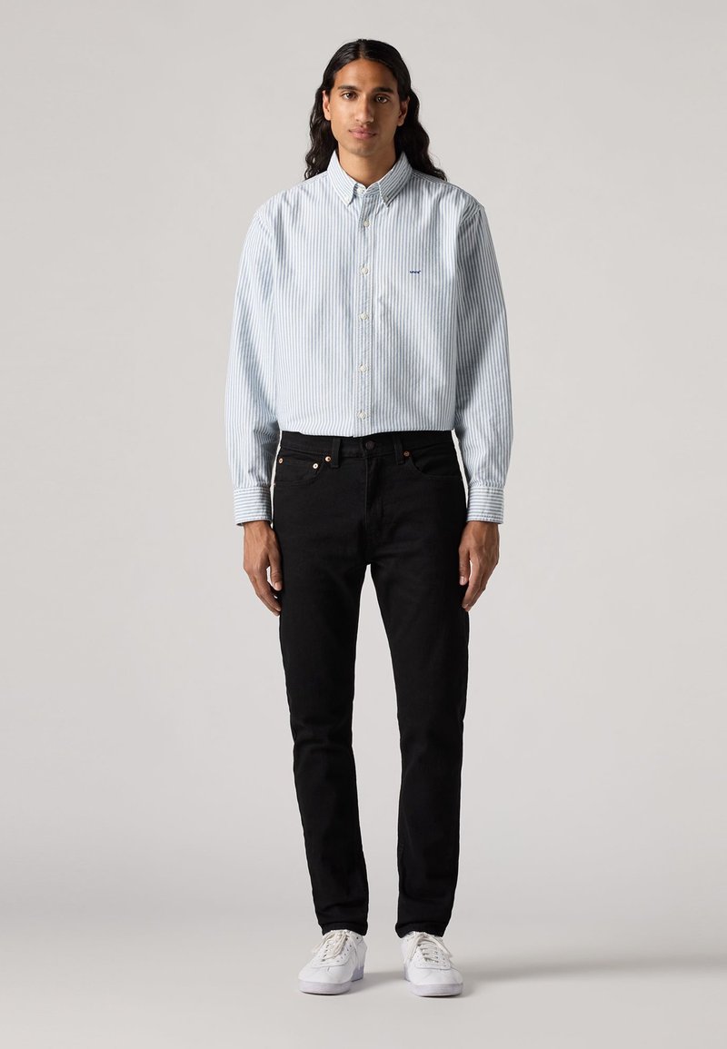Chemise boutonnée rayée bleu clair et blanche, jeans noirs et baskets blanches. La chemise possède un col et un logo sur la poitrine. Tissu lisse.