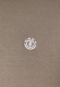 Tissu en coton kaki avec un logo blanc brodé centré sur la poitrine, présentant des éléments circulaires et linéaires dans son design.