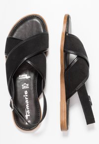 Black sandal featuring crisscrossed suede straps, a cushioned leather insole, and a rubber outsole with a brown accent. Adjustable heel strap.