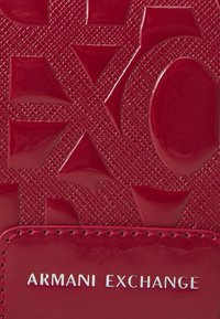 Glansig röd texturerad yta med olika geometriska former och ett sydd patch som visar "ARMANI EXCHANGE" i silverfärgade bokstäver.