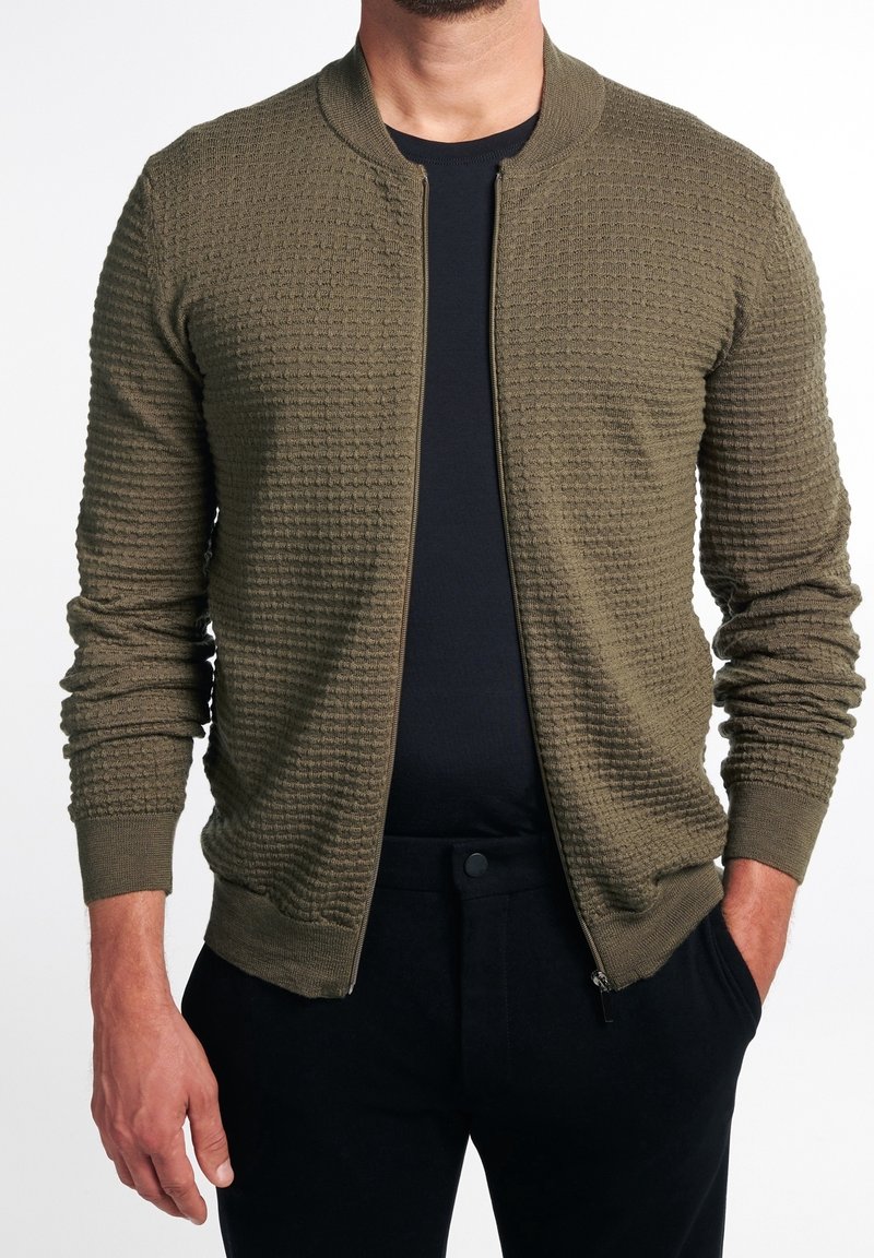 Homme portant un cardigan à fermeture éclair texturé vert olive sur une chemise noire et un pantalon noir, les mains détendues le long du corps.
