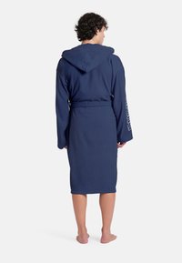 Accappatoio con cappuccio di colore blu navy realizzato in tessuto morbido e testurizzato; presenta maniche lunghe e una vita con cinturino, con un logo stampato sulla manica.