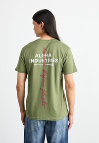Olivengrøn bomuld T-shirt med korte ærmer, med hvid og vinrød tekst på bagsiden, der siger "ALPHA INDUSTRIES" og "Rede before flight."