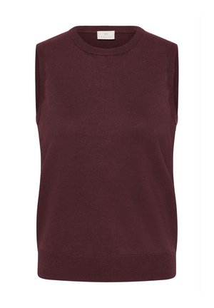 Femme debout, les mains dans les poches, portant un haut sans manches violet foncé, un pantalon large bleu foncé et des ballerines beiges sur un fond uni.