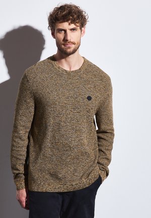Pull en tricot aux tons marron et jaune, col rond, manches longues, avec un patch logo noir sur la poitrine. Tissu texturé.