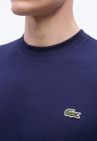 Άνδρας που φοράει μπλε ναυτικό μπλουζάκι με στρογγυλή λαιμόκοψη και μικρό πράσινο κεντημένο λογότυπο κροκόδειλου στο αριστερό στήθος.