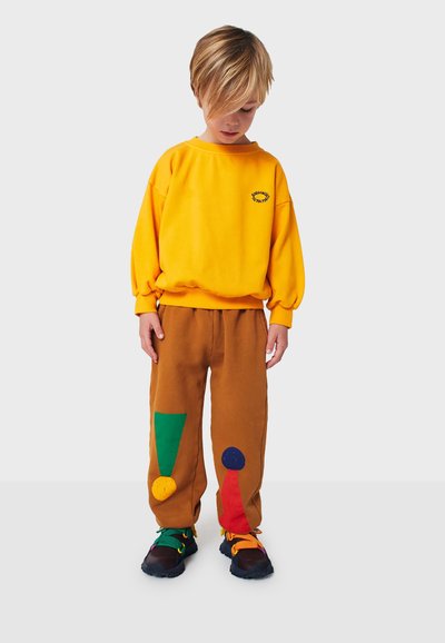 Felpa gialla con dettagli ricamati, pantaloni marroni con toppe colorate in verde, rosso e blu, abbinati a scarpe nere e colorate.