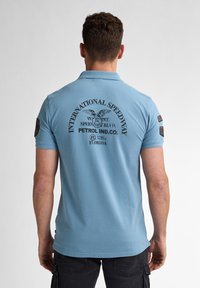 Ljusblå pikétröja med en svart tryckdesign på baksidan, inklusive "INTERNATIONAL SPEEDWAY", med patchdetaljer på ärmarna.