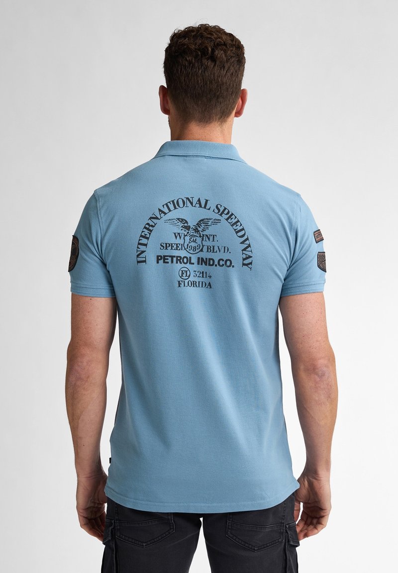 Ljusblå pikétröja med en svart tryckdesign på baksidan, inklusive "INTERNATIONAL SPEEDWAY", med patchdetaljer på ärmarna.