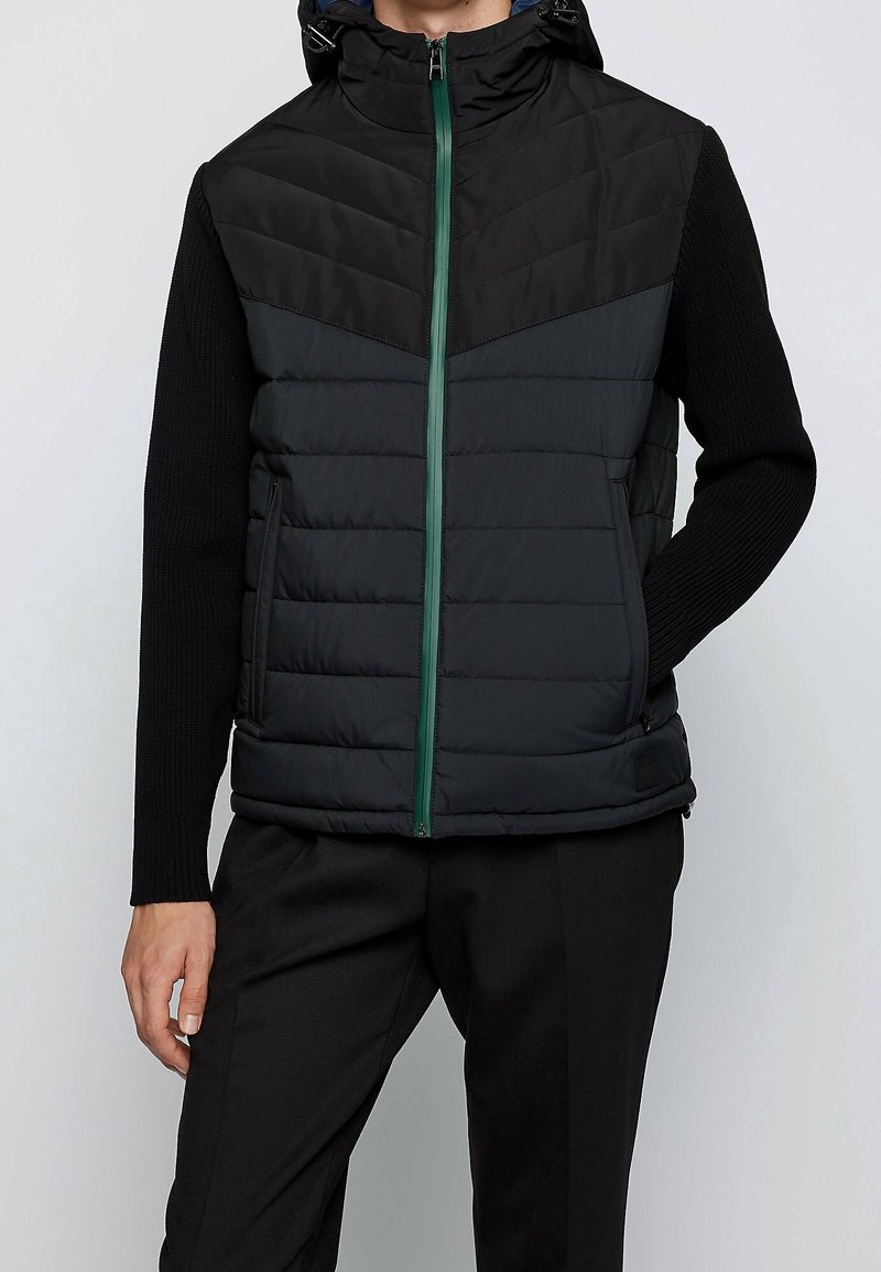 Personne portant une veste matelassée noire à capuche avec une fermeture éclair verte, par-dessus un pull noir à manches longues et un pantalon noir, debout avec une main dans la poche.