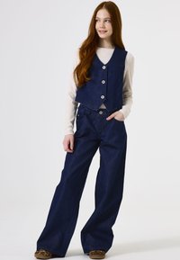 Navy denim vest met drie knopen, gecombineerd met een brede navy jeans. Lichte langslavige top zichtbaar onder de vest.