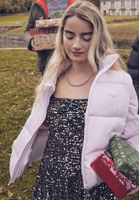 Luchtige lichtroze jas over een zwarte jurk met pailletten, met een kubusvormige silhouet. Cadeaus met inpakpapier in verschillende kleuren in de handen; groen en rood patroon.