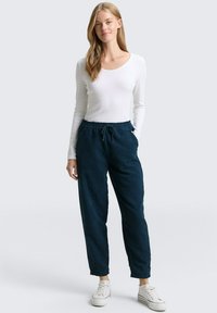 Marineblauwe korduroy broek met een elastische tailleband en trekkoorddetails, gecombineerd met een aangesloten witte lange mouwtop en witte sneakers.