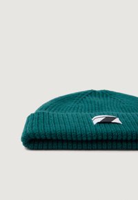 Gorro de punto en color verde azulado con una textura acanalada, que presenta un borde doblado y un pequeño parche de logo en blanco y negro en la parte frontal.