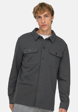 Jeune homme portant une veste-chemise gris foncé à boutons avec poches à rabat à l'avant, sur une chemise blanche, debout devant un fond uni.