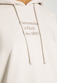 Hellbeiger Kapuzenpullover mit Kordelzug an der Kapuze. Mit braunem Aufdruck "Abercrombie & Fitch Since 1892" auf der Vorderseite. Weiches Material.