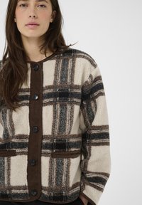 Fleecejacke mit einem cremefarbenen und braunen karierten Muster, versehen mit braunem Cordbesatz und schwarzen Knöpfen. Vorderseitige Taschen sind vorhanden.