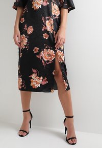 Černé květinové šaty s oranžovými a bílými květinami, saténová textura, střední délka, s bočním rozparkem a volným střihem. V páru s černými podpatky.