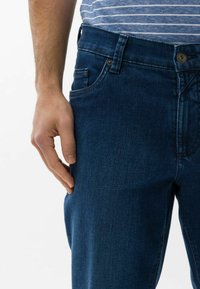 Jean en denim bleu foncé avec une texture lisse, coupe droite, style à cinq poches et fermeture à bouton à la taille.