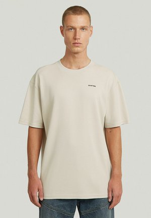Jonge man in een beige oversized G-Star T-shirt en blauwe spijkerbroek, staand tegen een effen lichtgrijze achtergrond, naar voren kijkend.