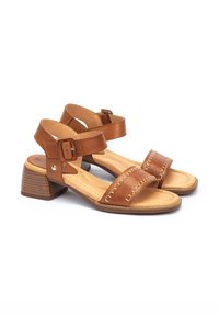 Sandalias de cuero marrón con una correa curva y hebilla en el tobillo, con un tacón texturizado y costuras decorativas en la parte delantera.