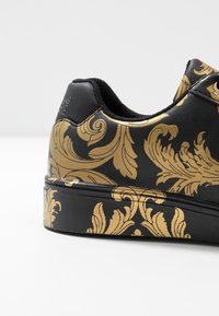 Černá teniska se zlatým barokním vzorem. Má texturovaný povrch a kontrastní černou podšívku. Plochá podrážka s kulatou špičkou.