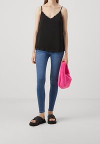 Camisole noire avec encolure festonnée, associée à un jean bleu ajusté et des sandales noires épaisses. Tient un sac à main duveteux rose vif.