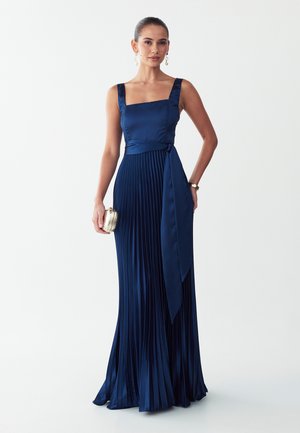 Vrouw die een mouwloze marineblauwe geplooide jurk met ceintuur draagt, een kleine gouden clutch vasthoudt en druppeloorbellen en een polshorloge draagt.