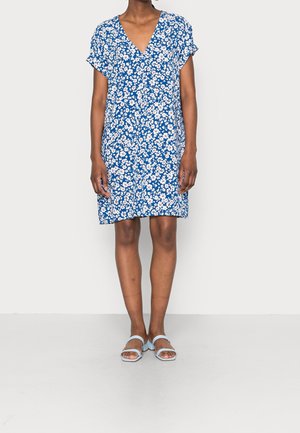 Femme portant une robe bleue à imprimé floral blanc et manches courtes, associée à des sandales bleu clair à bout ouvert, debout sur un fond uni.