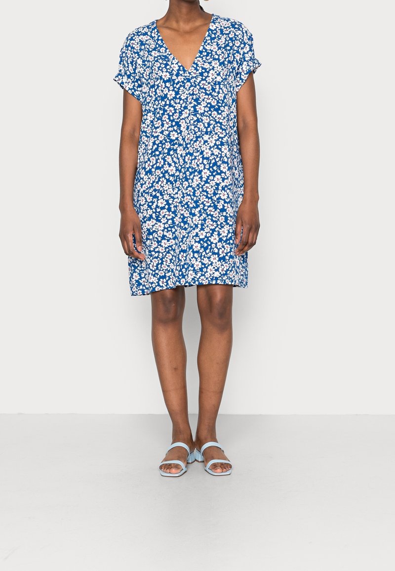 Femme portant une robe bleue à imprimé floral blanc et manches courtes, associée à des sandales bleu clair à bout ouvert, debout sur un fond uni.