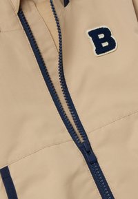 Beige jas met een marineblauwe rits, contrasterende afwerking en een geborduurde "B" patch op de borst. Hoge kwaliteit textuur en gestroomlijnd ontwerp.