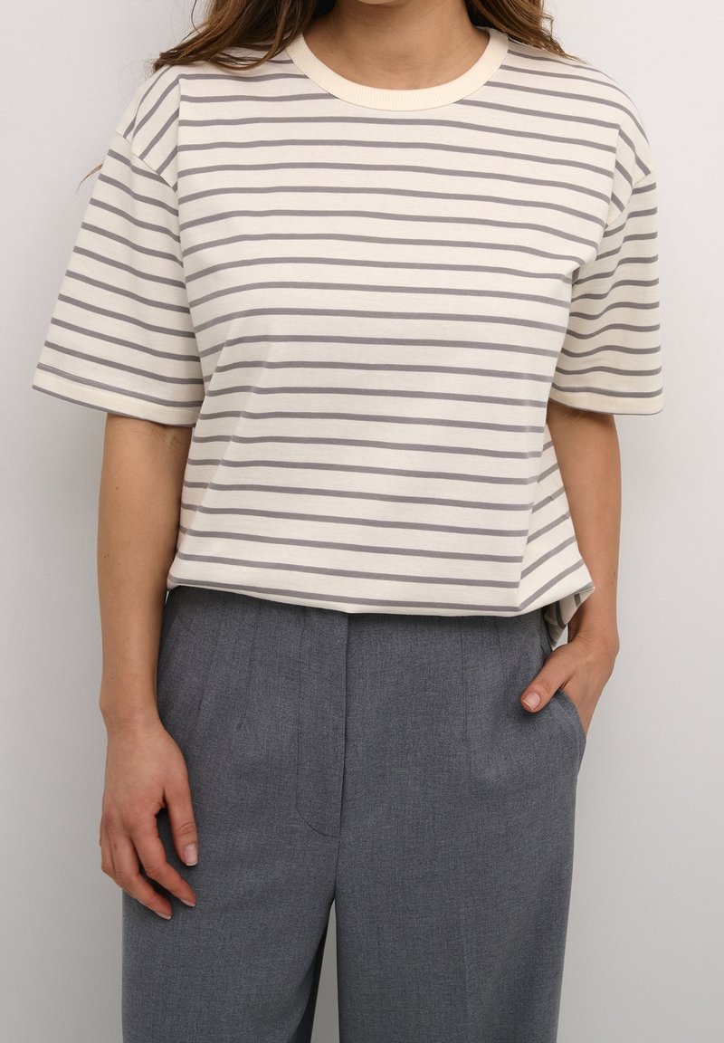 Femme portant une chemise à rayures crème et grises à manches courtes rentrée dans un pantalon gris taille haute, avec une main dans la poche.