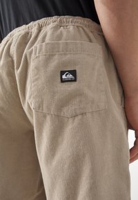 Short en velours côtelé beige avec une ceinture élastique, comportant une poche arrière et une petite étiquette noire avec logo. La texture est côtelée et douce.