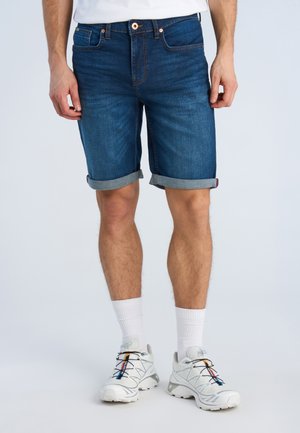 Mężczyzna w ciemnoniebieskich podwiniętych szortach dżinsowych, białych skarpetkach i białych sportowych butach z wielokolorowymi detalami, stojący na jasnym tle.