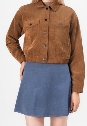 Person iført en brun kort kordsjakke med sorte knapper og et blåt kords A-formet skørt.