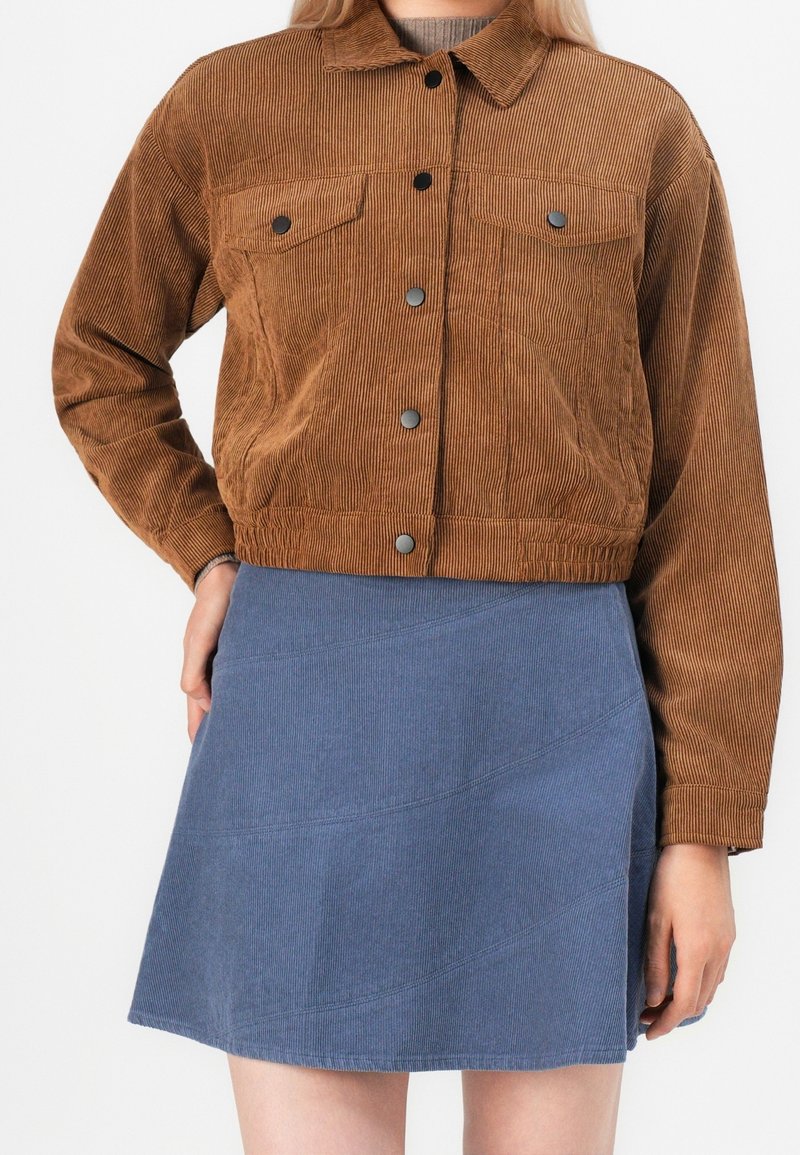 Personne portant une veste courte en velours côtelé marron avec des boutons noirs et une jupe trapèze en velours côtelé bleu.