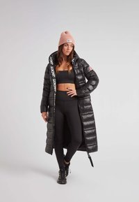 Mujer que lleva un abrigo de plumas largo negro, ropa deportiva negra, un gorro rosa y zapatos de senderismo, de pie contra un fondo gris claro.