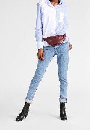 Personne portant une chemise bicolore bleu et blanc, un jean bleu clair retroussé, des bottines noires et une sacoche en cuir marron à la taille.