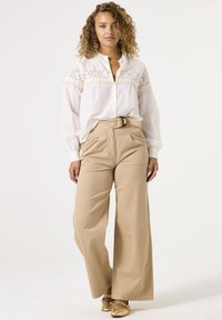 Weißes Hemd mit Knöpfen und stickereien, kombiniert mit beigen, weit geschnittenen Hosen. Goldene Schuhe runden das Outfit ab. Glatte Stofftextur.