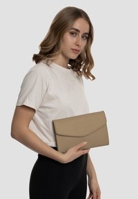 Clutch in pelle beige con finitura testurizzata, caratterizzato da un design ripiegato e un accento in logo metallico, tenuto da un modello in abbigliamento casual.