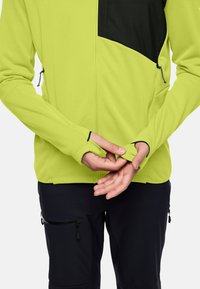 Giacca verde lime con zip e pannello nero sulle spalle, dotata di fori per i pollici, tasche con zip e un tessuto leggero e liscio.