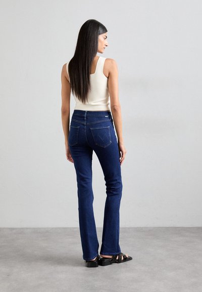 Dark blue flared jeans with a high waist and rear pockets, worn with a cream tank top. The model has straight hair and black sandals.