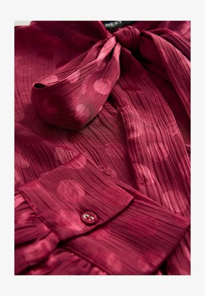 Blouse bourgogne en tissu texturé, avec un col noué, des plis verticaux et de grands motifs à pois ; comprend un détail de poignet avec boutons.