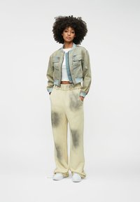 Νεαρή γυναίκα στέκεται με τα χέρια στις τσέπες, φοράει ένα κοντό τζιν μπουφάν, λευκό κοντό μπλουζάκι, κρεμ παντελόνι τύπου tie-dye με φαρδιά γραμμή και λευκά αθλητικά παπούτσια.