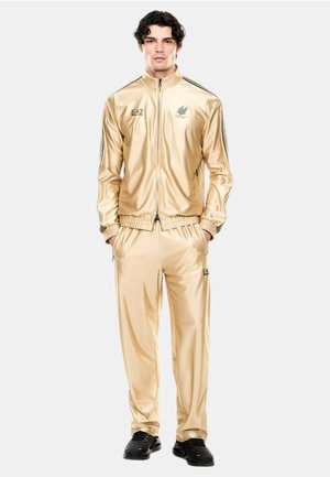 Uomo in piedi indossa una tuta beige lucida con giacca con zip e pantaloni abbinati, abbinata a sneaker nere, mani nelle tasche, su sfondo bianco.