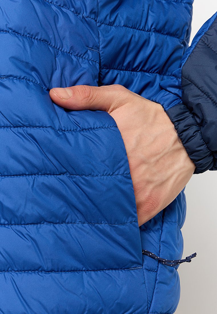 Chaqueta de abrigo azul con textura acolchada, puños elásticos y un bolsillo visible con cordón, que muestra un material suave y brillante.