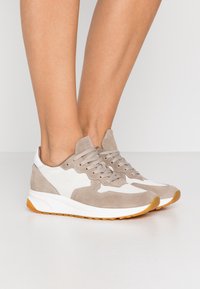 Jalat memakai sepatu sneaker suede beige dan putih dengan sol karet gum, berdiri di atas lantai putih di latar belakang putih polos.