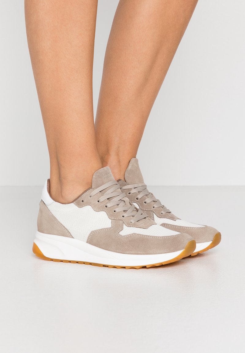 Jalat memakai sepatu sneaker suede beige dan putih dengan sol karet gum, berdiri di atas lantai putih di latar belakang putih polos.