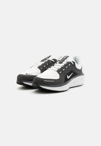 Zapatos deportivos en blanco y negro con una parte superior texturizada, lengüeta acolchada y lazadas planas, que presentan un pequeño logotipo y una suela de goma para mayor tracción.